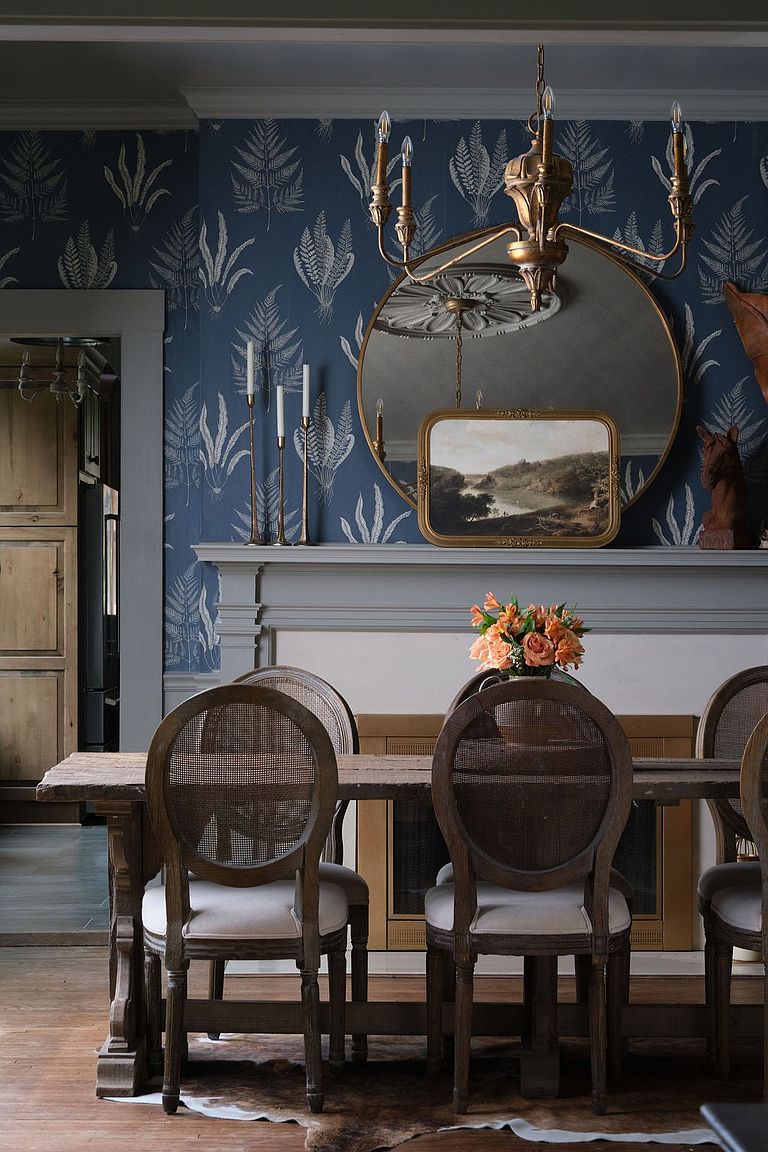 This is an interior shot of a dining room featuring a rustic wooden table surrounded by chairs with woven backs and light-colored cushions. A round mirror and a framed painting hang above a gray fireplace mantel, complemented by a gold chandelier. The walls are adorned with a blue botanical wallpaper, creating a sophisticated and inviting atmosphere.