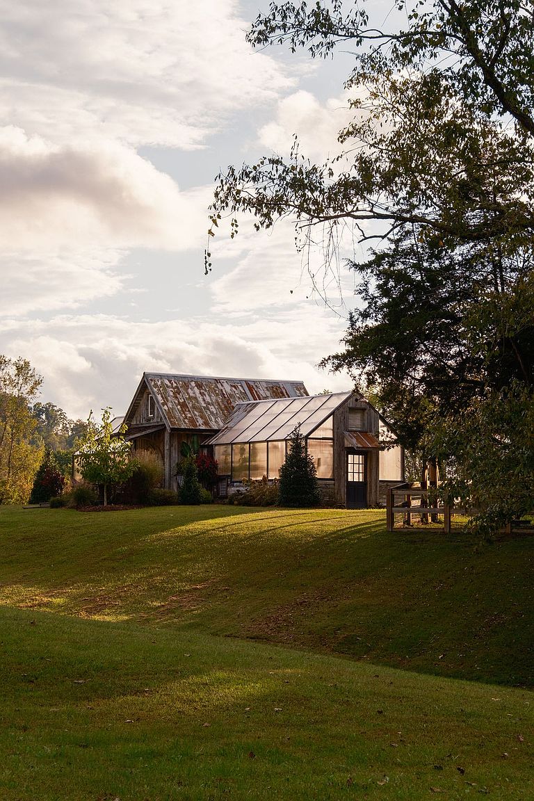 This image showcases a charming property with a well-maintained lawn and a rustic barn-style structure connected to a greenhouse. The building features a weathered roof and wooden siding, blending seamlessly with the natural surroundings. The scene is bathed in soft, natural light, creating a warm and inviting atmosphere.