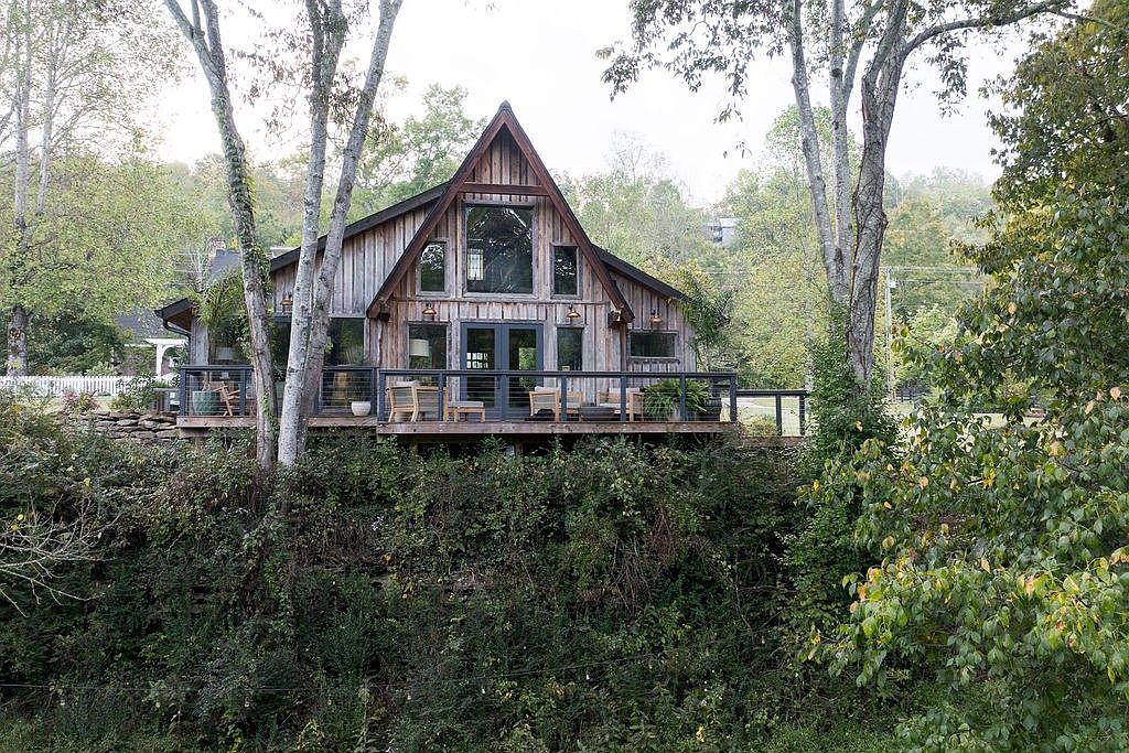 This is a front view of a rustic, A-frame house with a large deck. The house is nestled among trees and greenery, giving it a secluded and natural feel. The deck features wooden furniture and potted plants, enhancing the outdoor living space.