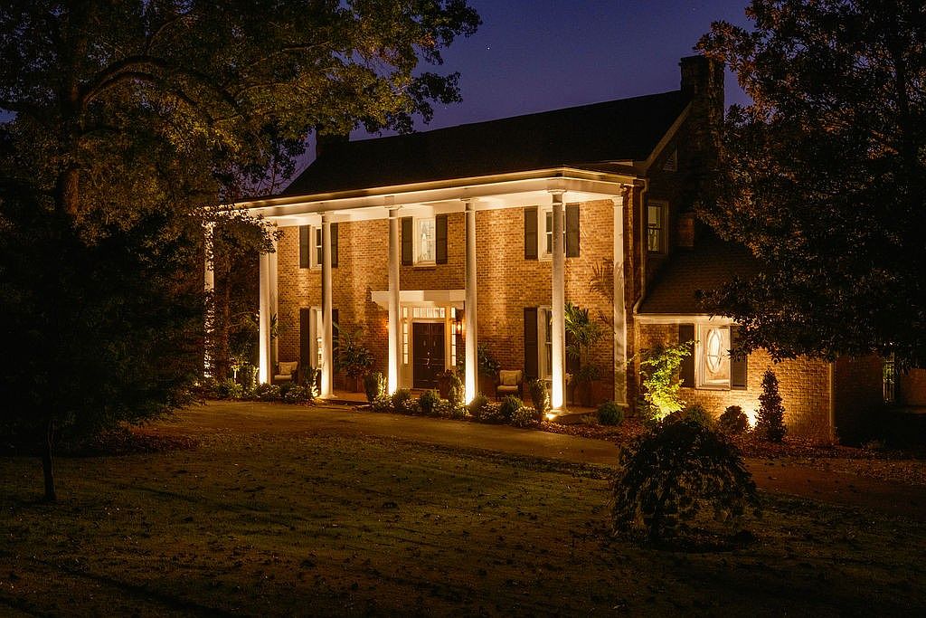 This is a front exterior view of a grand, two-story brick house at night, illuminated by landscape lighting. The house features a prominent portico with white columns, dark shutters, and a symmetrical facade. The well-manicured lawn and mature trees add to the property's curb appeal, creating an inviting and stately impression.