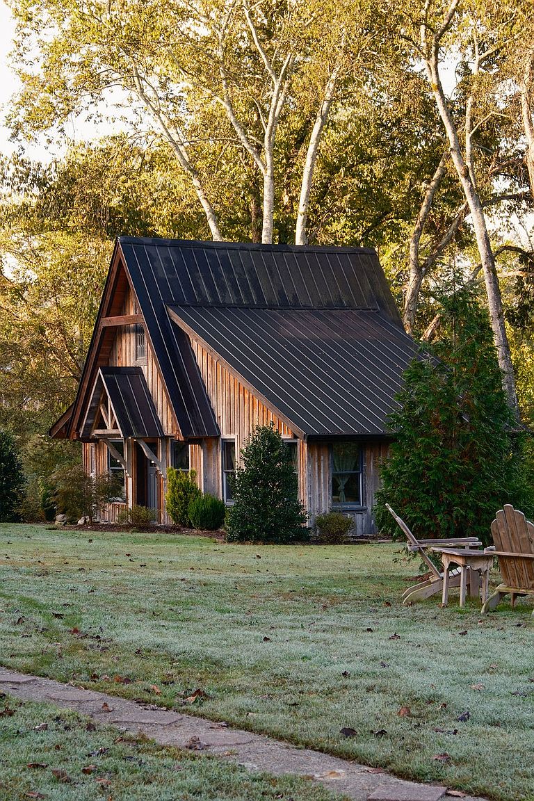 This image showcases the front exterior of a charming A-frame cabin nestled among trees. The cabin features a dark metal roof, natural wood siding, and a small covered entryway. The surrounding yard is covered in grass, with a stone pathway leading to the cabin and wooden chairs visible on the right, creating a cozy and inviting atmosphere.