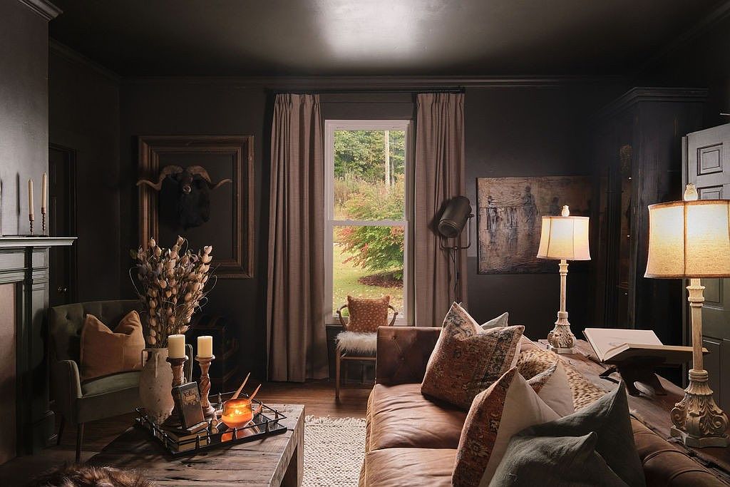 This is a cozy and sophisticated living room with dark walls and warm lighting. A leather sofa is adorned with decorative pillows, and a rustic coffee table displays candles and decorative items. The room features a window with curtains, artwork, and lamps, creating a comfortable and inviting atmosphere.
