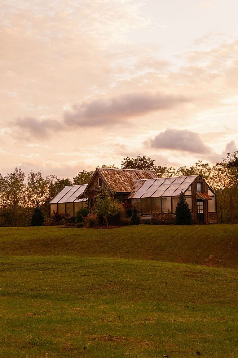 This image showcases a charming greenhouse nestled on a lush green lawn, set against a backdrop of trees and a soft, cloudy sky. The greenhouse features a rustic design with a weathered roof and glass panels, creating an inviting and serene atmosphere. The scene evokes a sense of tranquility and natural beauty, highlighting the property's potential for gardening and outdoor enjoyment.
