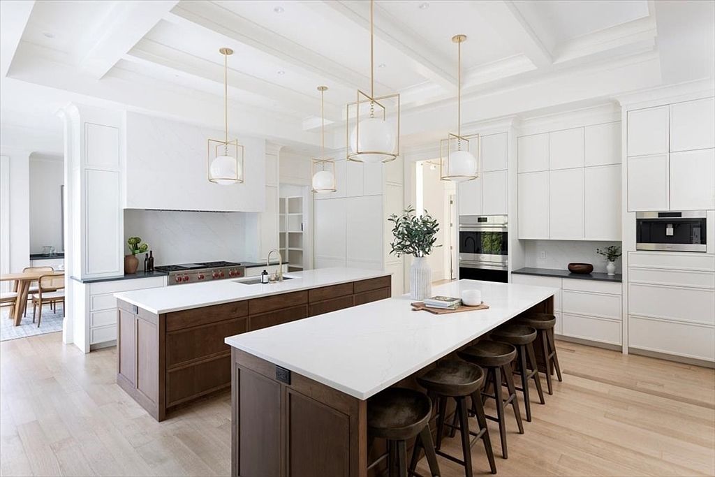 This expansive, bright kitchen features a dual-island layout with dark wood cabinetry contrasted against crisp white perimeter cabinets and stone countertops. The space is illuminated by elegant pendant lighting and includes high-end built-in appliances, creating a sophisticated and functional culinary environment. The light wood flooring and coffered ceiling details add warmth and architectural interest to this modern, open-concept design.