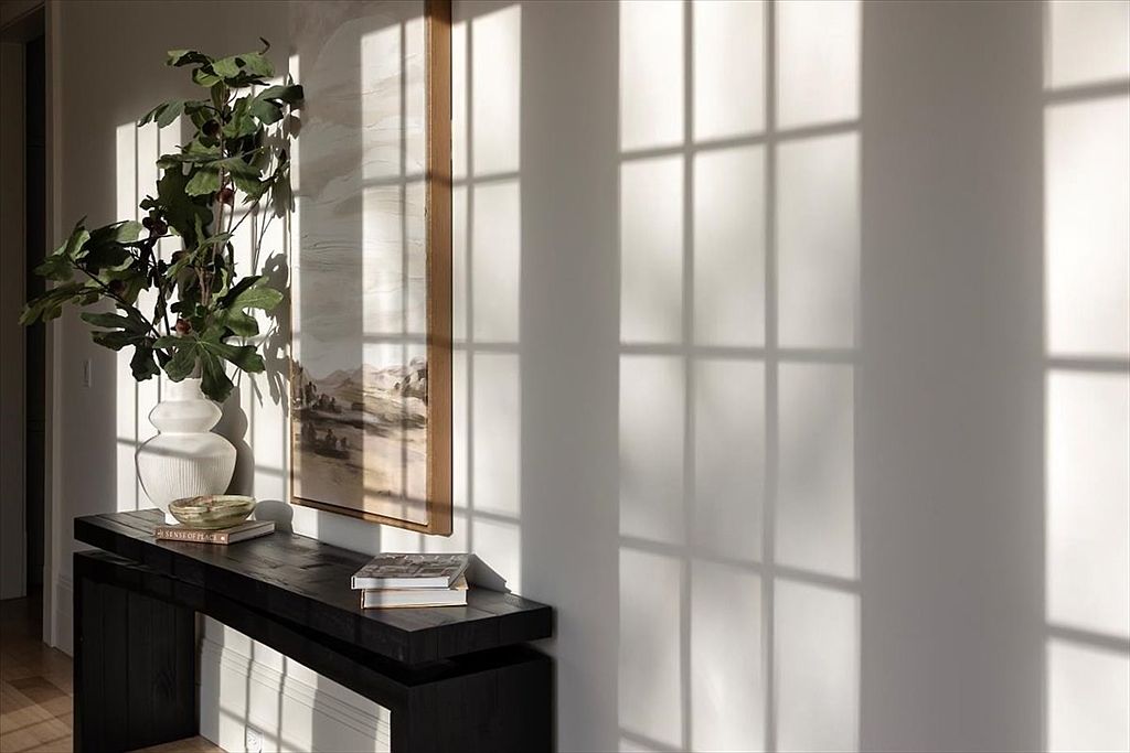 This interior vignette features a dark, minimalist console table positioned against a neutral wall, adorned with a decorative vase of greenery and a stack of books. A framed landscape artwork hangs above the table, while dramatic, grid-patterned shadows cast by nearby windows create a striking geometric effect across the wall. The composition offers a serene, sophisticated aesthetic, highlighting the interplay of light and shadow in a transitional space.