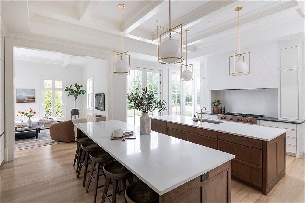 This bright, open-concept kitchen features dual islands with light-toned stone countertops and dark wood cabinetry, creating a sophisticated contrast. The space is illuminated by elegant gold-toned pendant lights and features a large, clean-lined range hood above a professional-style stove. A seamless transition leads into an adjacent living area, emphasizing the airy, high-end design of this modern home.