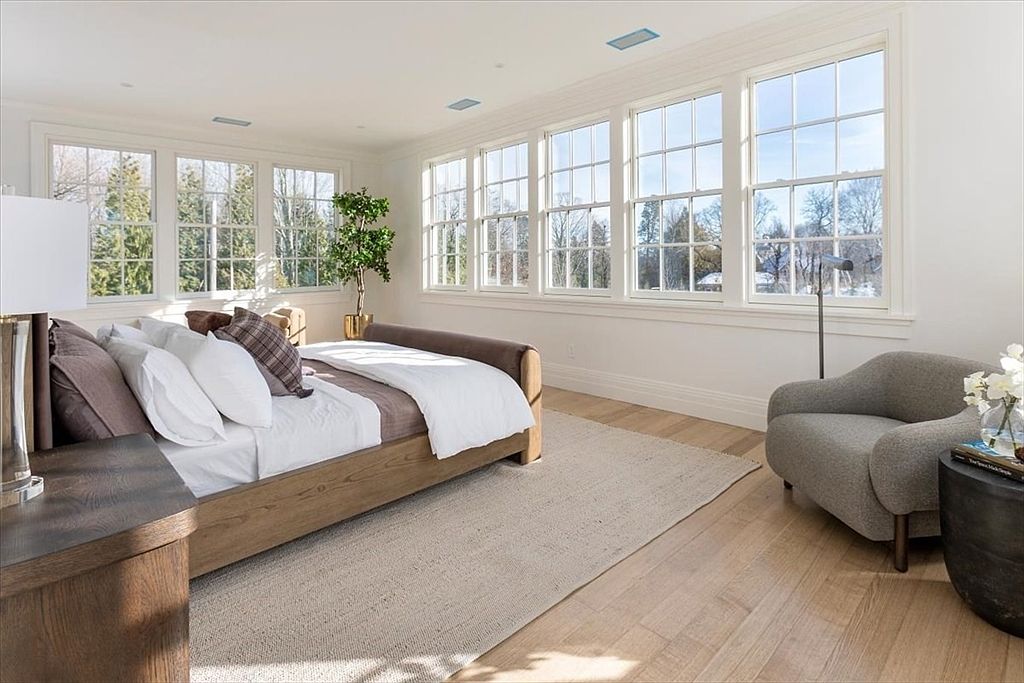 This bright and airy primary bedroom features light wood flooring and a neutral color palette, centered around a wooden bed frame with plush bedding. A wall of large, multi-pane windows allows for abundant natural light and views of the surrounding trees, while a cozy gray armchair sits in the corner. The room is accented by a potted plant and a dark side table, creating a serene and sophisticated atmosphere.