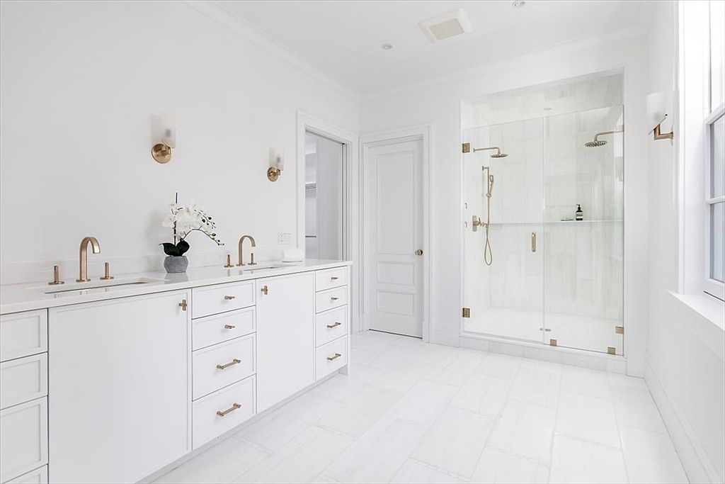 This bright and airy primary bathroom features a clean, all-white aesthetic with a long vanity topped with stone counters and gold-toned fixtures. A glass-enclosed walk-in shower with light-colored tile occupies the corner, while the room is finished with matching white floor tiles. The perspective is a wide-angle shot from the doorway, showcasing the minimalist design and high-end finishes.
