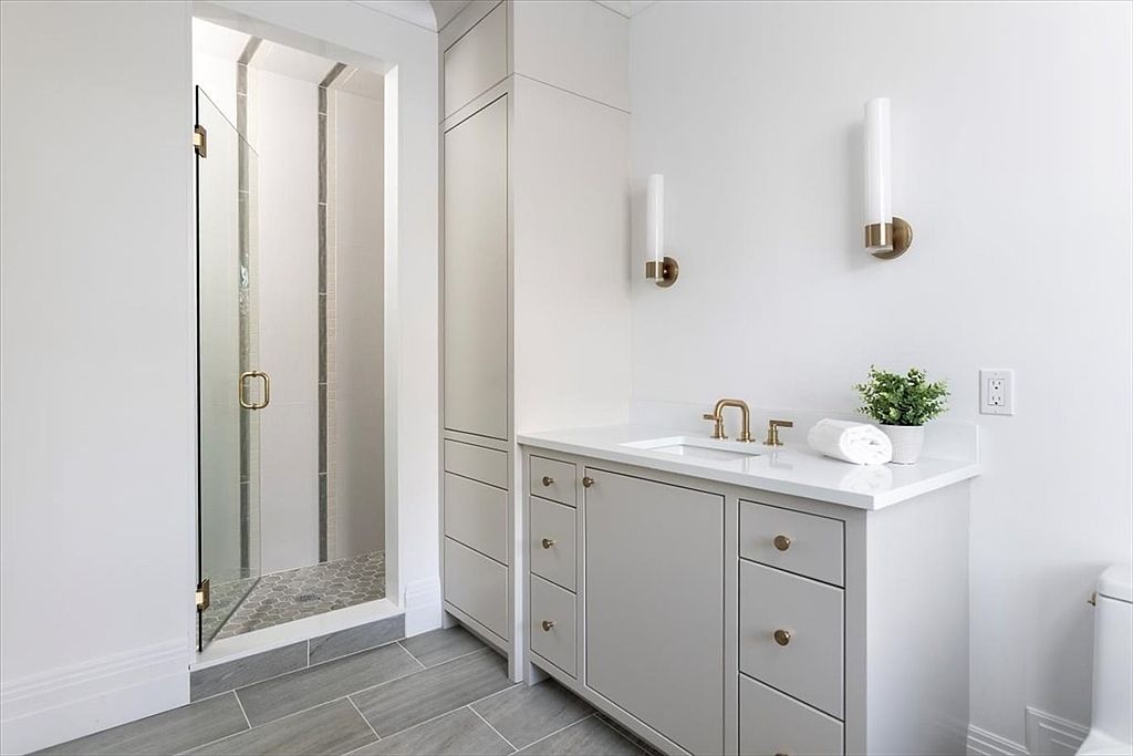 This bright and modern bathroom features a light gray vanity with a white countertop and gold-toned hardware. To the left, a glass-enclosed shower showcases patterned tile flooring, while two wall-mounted light fixtures flank the vanity mirror area. The space is finished with neutral wall tones and gray wood-look floor tiles, creating a clean and cohesive aesthetic.
