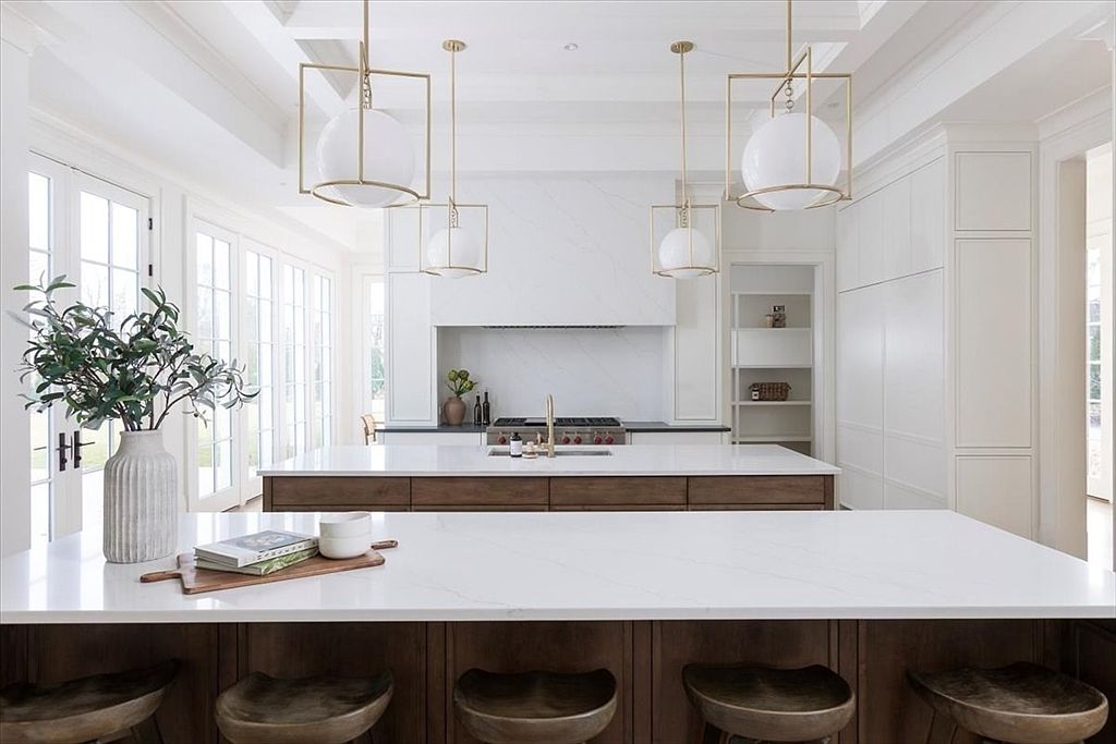 This bright and airy kitchen features a dual-island layout with dark wood cabinetry topped by light-toned stone countertops. Four geometric pendant lights hang above the islands, while a professional-style range is set into a clean, white-clad alcove. The space is accented by a decorative vase with greenery and bar-style seating, creating a sophisticated and functional culinary environment.