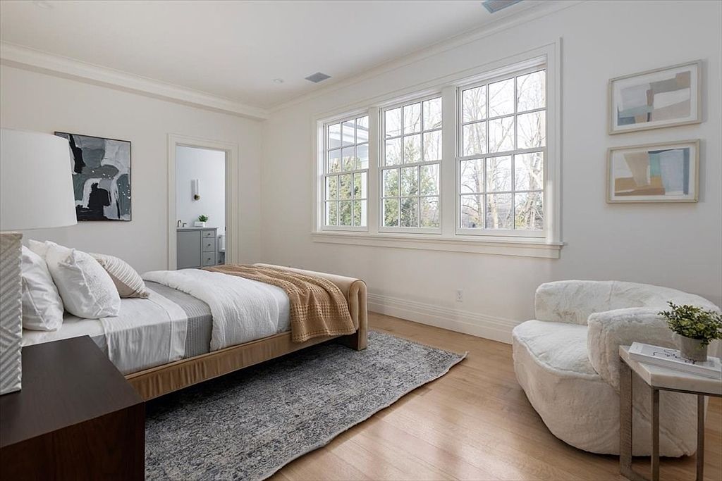 This bright and airy bedroom features light wood flooring, neutral walls, and a large window arrangement that fills the space with natural light. The room is furnished with a bed dressed in white linens with a tan throw, a textured area rug, and a plush white armchair positioned near a small side table. A doorway leads to an adjacent bathroom, and the overall aesthetic is clean, modern, and inviting.