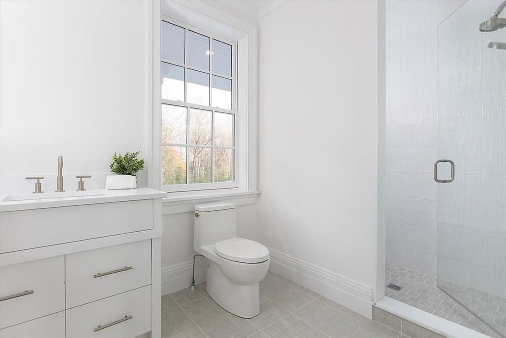 This bright and clean bathroom features a white vanity with a stone countertop, a modern toilet, and a glass-enclosed shower with light-colored tile. A large, multi-pane window allows natural light to fill the space, creating an airy and inviting atmosphere. The neutral color palette and minimalist design provide a contemporary and well-maintained aesthetic.