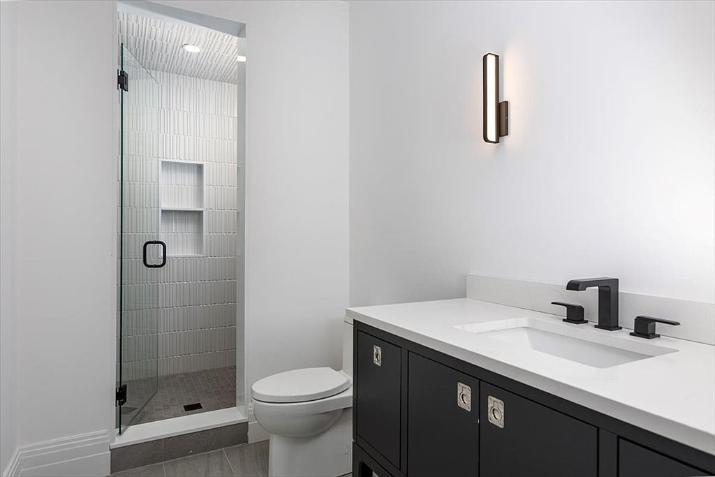 This modern bathroom features a sleek, dark vanity with a white countertop and matte black fixtures. To the left, a glass-enclosed shower stall showcases textured white wall tiles and a built-in recessed niche. The space is bright and minimalist, highlighted by a contemporary wall-mounted light fixture above the vanity.