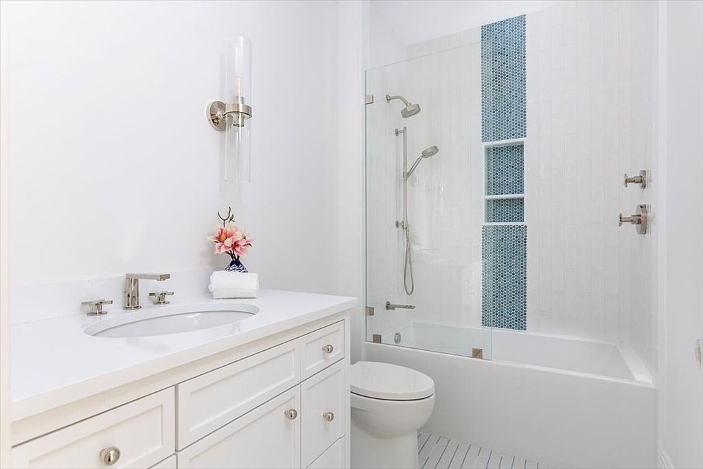 This bright and clean bathroom features a white vanity with a stone countertop and a single undermount sink. A glass-enclosed shower-tub combination is accented by a vertical strip of blue mosaic tiles and built-in shelving. The space is illuminated by a sleek wall-mounted light fixture, creating a crisp and modern aesthetic.