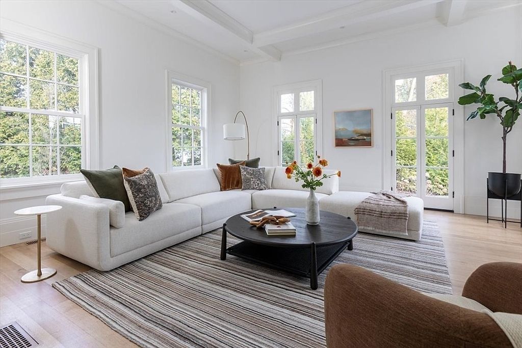This bright and airy living room features a large, neutral-toned sectional sofa arranged around a dark circular coffee table on a textured, striped area rug. The space is filled with natural light from multiple tall windows and glass doors that look out onto a lush, green exterior. A tall potted plant adds a touch of nature to the corner, while minimalist decor and soft textiles create a welcoming, contemporary atmosphere.