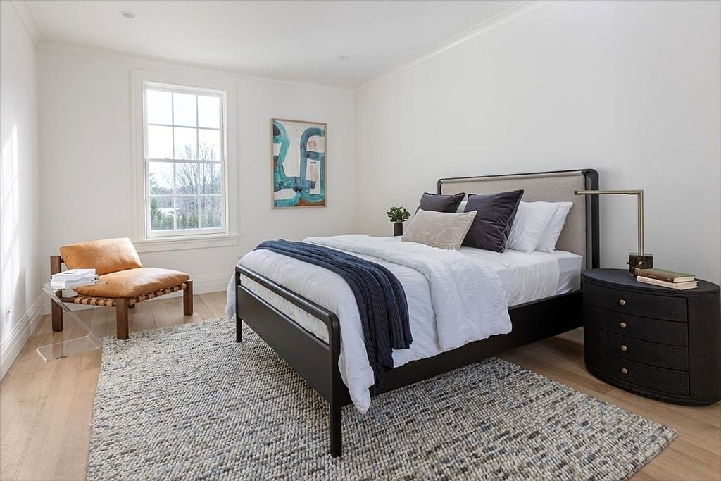 This bright and airy bedroom features a modern metal-framed bed dressed in crisp white linens and a navy throw, centered against a neutral wall. To the left, a tan leather lounge chair sits on a clear acrylic base near a large window, while a dark, curved nightstand with a sleek lamp anchors the right side of the bed. A textured, multi-toned area rug covers the light wood flooring, and a vibrant abstract piece of art adds a pop of color to the minimalist space.