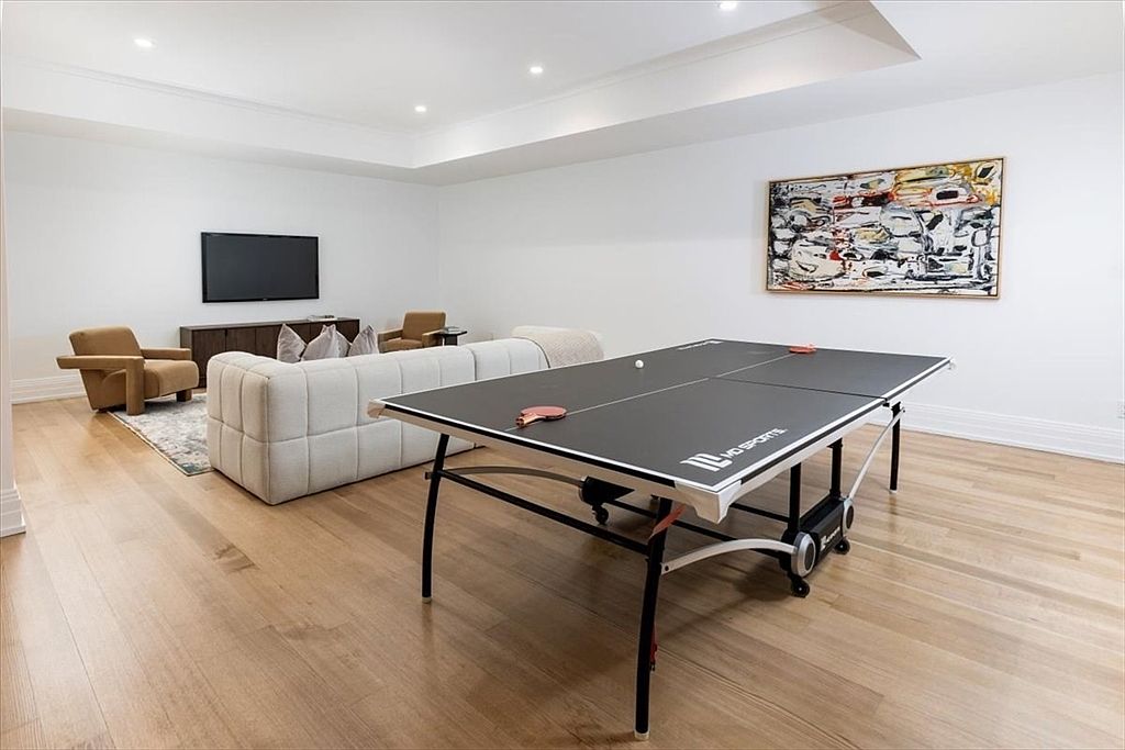 This spacious, open-concept recreation room features light wood flooring and bright white walls, creating a clean and versatile living area. The space is furnished with a comfortable light-colored sofa and two armchairs facing a wall-mounted television, while a ping-pong table occupies the foreground. A large, colorful abstract painting adds a vibrant focal point to the room, which is illuminated by recessed ceiling lighting.