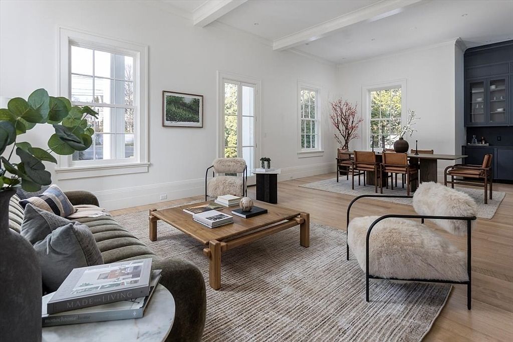 This bright and airy open-concept living and dining area features light wood flooring and crisp white walls, creating a sophisticated, modern aesthetic. The living space is anchored by a textured area rug, a low-profile sofa, and two plush, fur-accented armchairs, while the adjacent dining area is furnished with a long wooden table and leather-upholstered chairs. Large windows throughout the space provide ample natural light, and a dark-toned built-in cabinetry unit adds a striking contrast to the neutral palette.