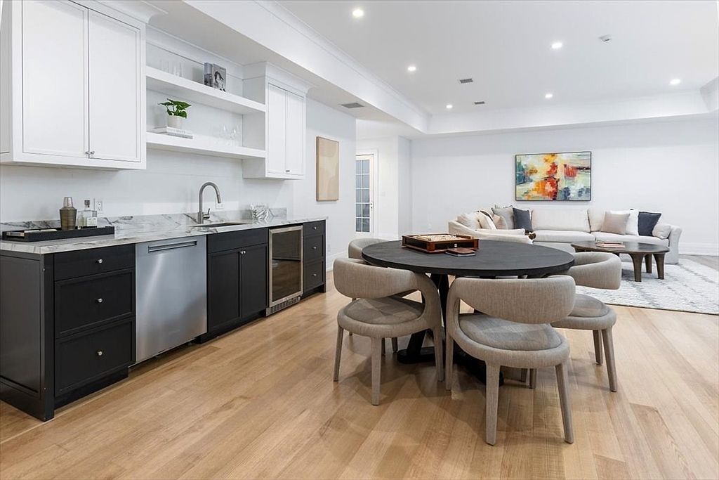 This open-concept space features a wet bar area with dark cabinetry, a stainless steel dishwasher, and a wine cooler, all topped with light-toned stone counters. A circular dark wood dining table with four upholstered chairs sits in the foreground, while a bright living area with a white sofa and abstract wall art is visible in the background. The room is finished with light wood flooring and recessed lighting, creating a clean, modern, and inviting atmosphere.