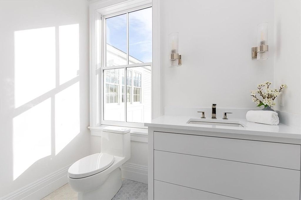 This bright and clean bathroom features a white vanity with a stone countertop and a modern undermount sink. A tall window allows natural light to fill the space, casting soft shadows on the wall, while a white toilet sits adjacent to the vanity. The room is accented with minimalist wall sconces and a small decorative floral arrangement, creating a fresh and airy atmosphere.