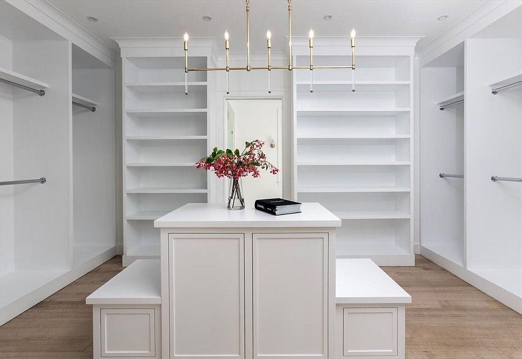 This elegant walk-in closet features crisp white cabinetry, extensive open shelving, and hanging rods arranged symmetrically on either side. A central island with a smooth top provides a focal point, complemented by two built-in benches and a sophisticated gold-toned chandelier hanging above. The light wood flooring adds warmth to the clean, minimalist aesthetic of this organized storage space.