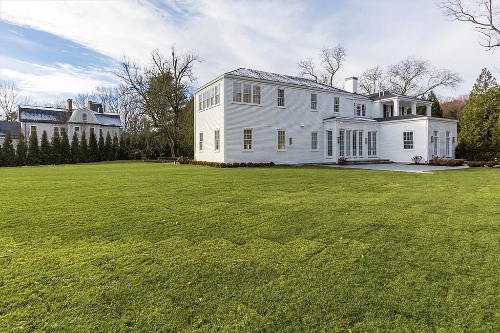 This rear view of a large, white-sided colonial-style home showcases a sprawling, well-manicured lawn that extends from the foreground toward the structure. The house features a symmetrical design with numerous multi-pane windows, a stone patio area, and a second-story balcony. A neighboring historic-style home is visible in the background, framed by mature trees under a bright, partly cloudy sky.