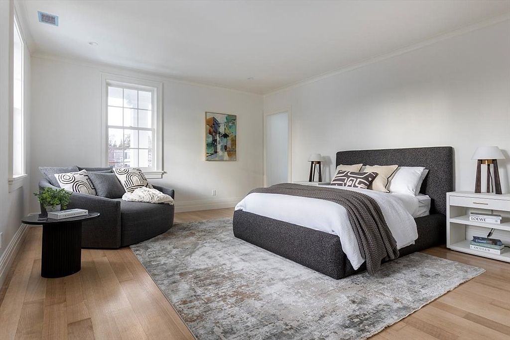 This bright and airy primary bedroom features a neutral color palette with a large, dark upholstered bed as the focal point. To the left, a cozy, rounded armchair sits near a window, creating a comfortable reading nook, while a white nightstand with open shelving provides storage. The room is finished with light wood flooring and a large, textured area rug, offering a clean and sophisticated aesthetic.