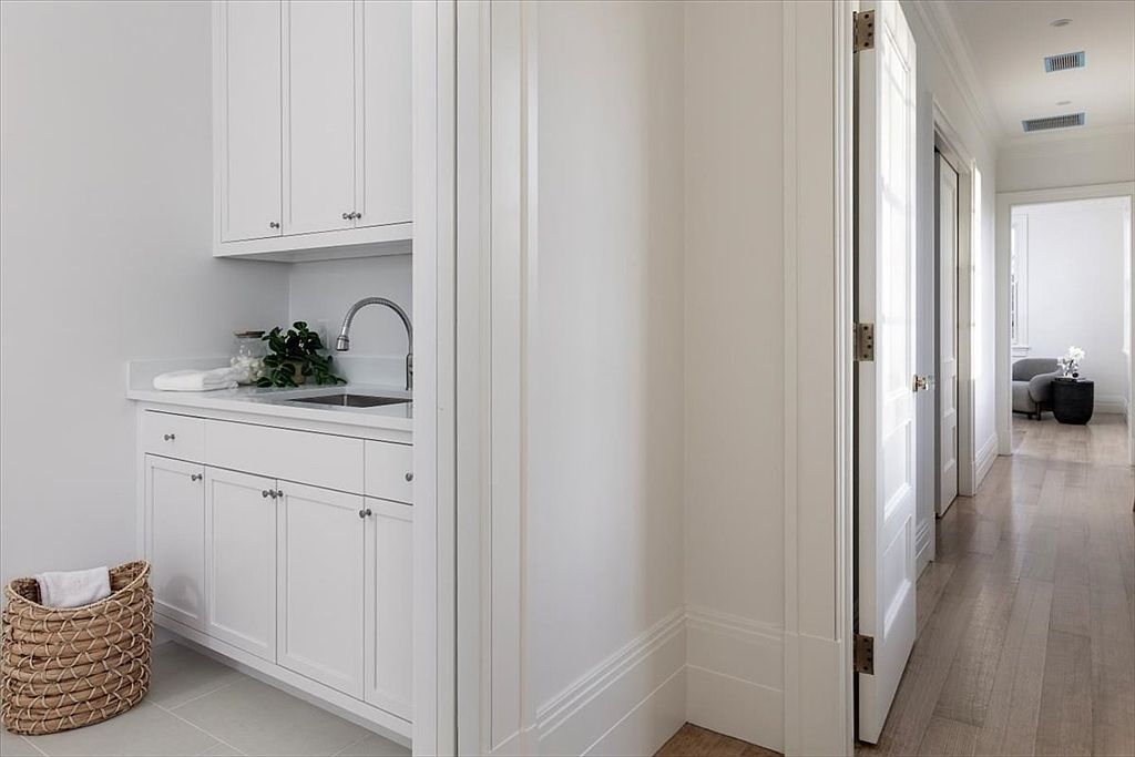 This bright and clean laundry area features white cabinetry with a built-in sink and a stone countertop. A woven basket sits on the light-colored tile floor, while a doorway leads into a hallway with light wood flooring. The space is minimalist and functional, showcasing a seamless transition between the utility area and the rest of the home.