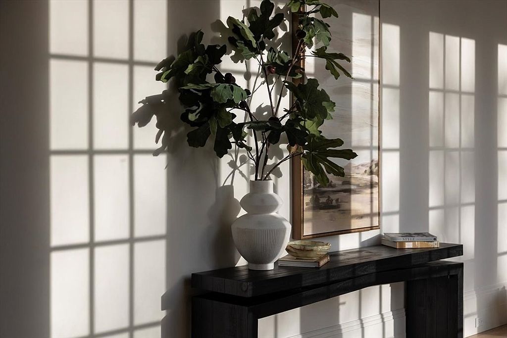 This elegant vignette features a dark, textured console table positioned against a neutral wall, serving as a sophisticated focal point. A sculptural white vase filled with lush greenery sits atop the table, complemented by a framed landscape artwork and a small bowl. The scene is artistically illuminated by dramatic, elongated shadows cast by nearby window panes, creating a serene and high-end atmosphere.