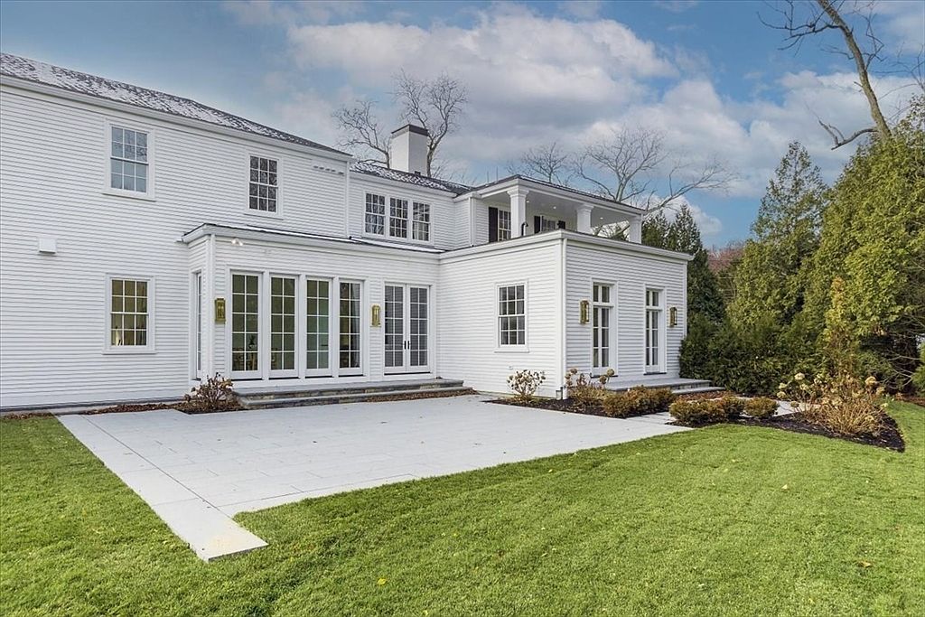 This rear view of a large, white-sided colonial-style home showcases a clean, symmetrical aesthetic with multiple multi-pane windows and a second-story balcony. A spacious, light-colored stone patio extends from the house, providing a transition to the well-manicured green lawn. The scene is framed by mature trees and a clear blue sky, creating a bright and inviting atmosphere.