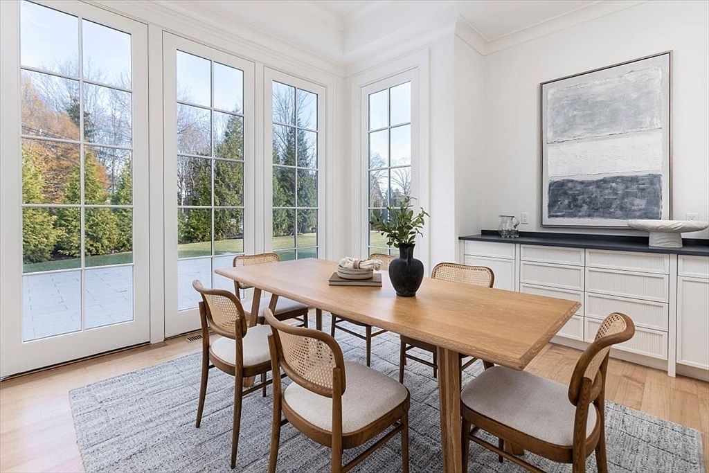 This bright dining area features a light wood table surrounded by four chairs with woven cane backs and neutral upholstered seats. A large, textured abstract painting hangs above a built-in sideboard with a dark top, while tall windows provide ample natural light and views of the outdoor greenery. The space is anchored by a light-colored area rug, creating a clean, modern, and inviting atmosphere.