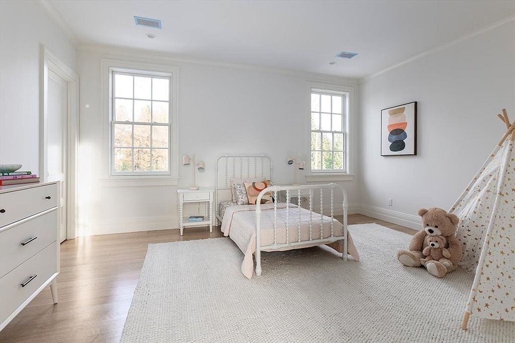 This bright and airy bedroom features a white metal-frame bed centered between two windows, creating a clean and inviting atmosphere. A soft, textured area rug covers the light wood flooring, while a playful teepee tent and a large teddy bear add a charming touch to the corner. The room is finished with a white dresser and minimalist decor, offering a peaceful and well-lit space.