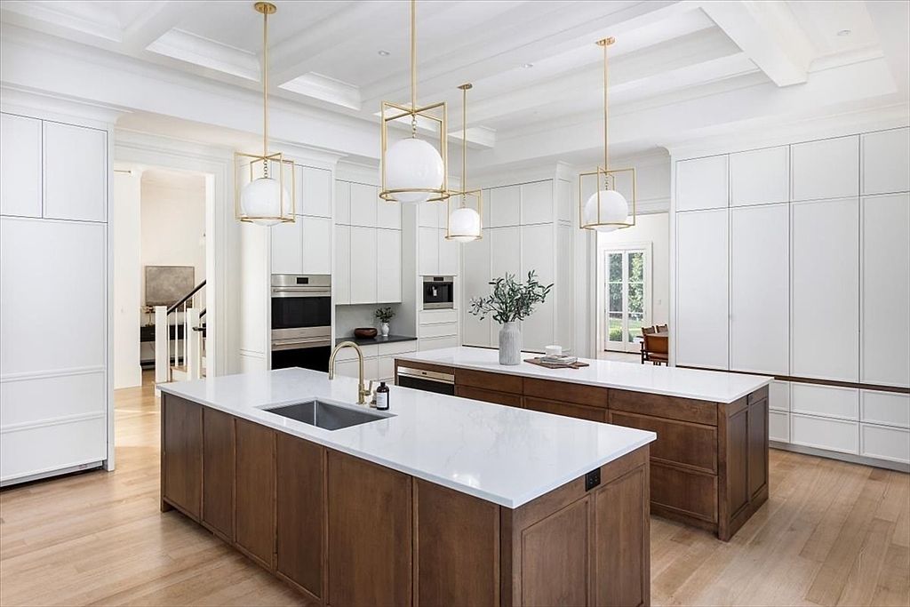 This spacious, high-end kitchen features a dual-island layout with rich-toned wood cabinetry and crisp white countertops. The room is illuminated by three elegant gold-framed pendant lights hanging from a coffered ceiling, while tall white cabinetry provides extensive storage along the perimeter. The perspective is a wide-angle shot from the kitchen floor, showcasing the clean lines, modern appliances, and the seamless transition to the adjacent hallway and dining area.