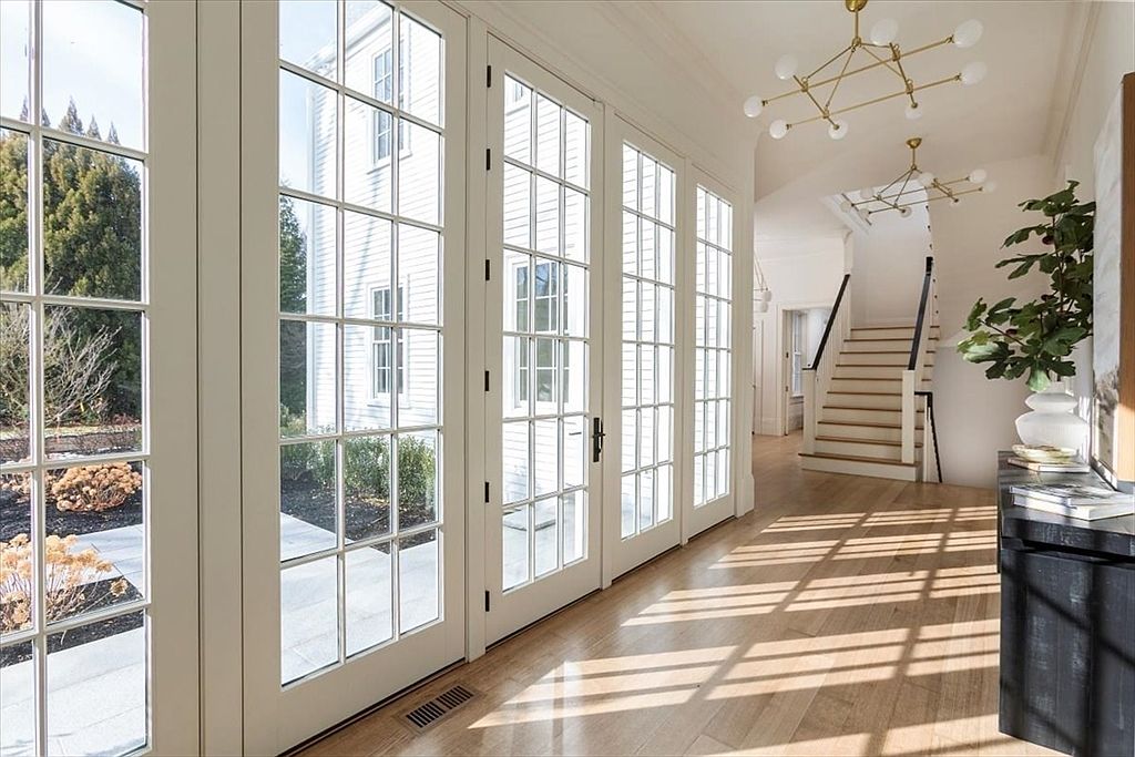 This bright and airy hallway features light wood flooring and a series of tall, multi-paned glass doors that allow abundant natural light to flood the space. A staircase with a dark railing leads to the upper level, while modern gold-toned chandeliers add a touch of elegance to the clean, white-walled interior. The perspective captures the transition from the sunlit entryway toward the staircase, highlighting the open and inviting atmosphere of the home.