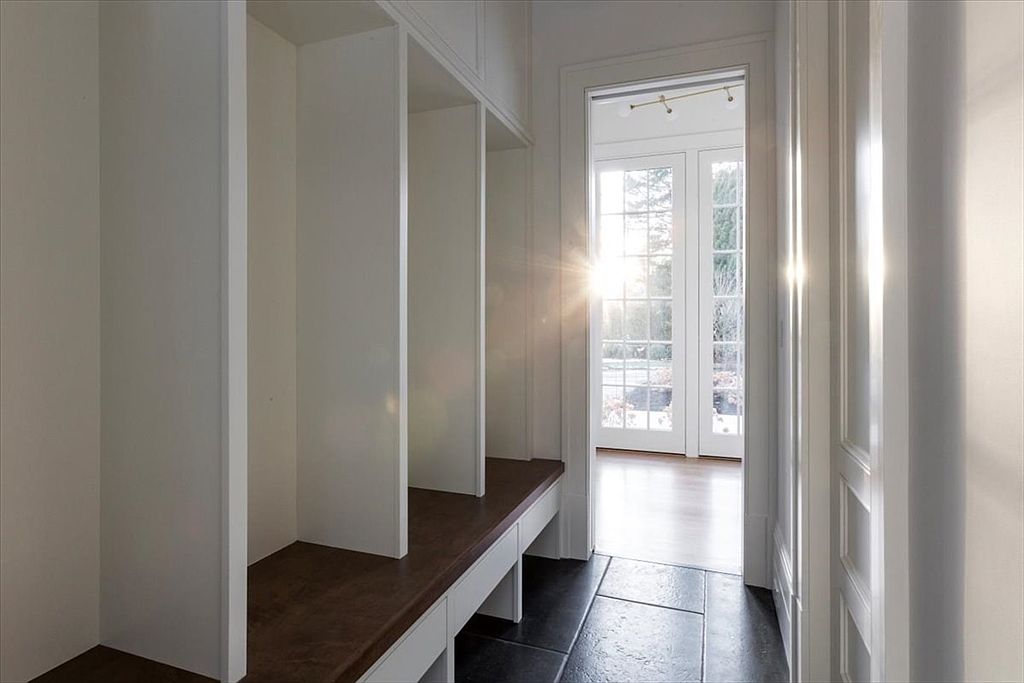 This bright mudroom or entryway transition space features built-in storage cubbies with a dark-toned bench seat. The area is finished with dark tile flooring and leads through a doorway toward a set of glass-paned double doors that allow natural light to flood the space. The clean, white-painted woodwork and minimalist design create a welcoming and functional atmosphere.