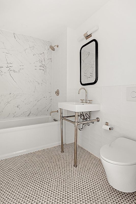 This is a well-lit bathroom featuring a modern design with a white marble-like tiled wall in the shower/tub area and white tiled walls elsewhere. The floor has a geometric pattern with small triangles. A sleek, wall-mounted sink with chrome legs and a black-framed mirror add to the contemporary aesthetic, while a white toilet sits to the right.