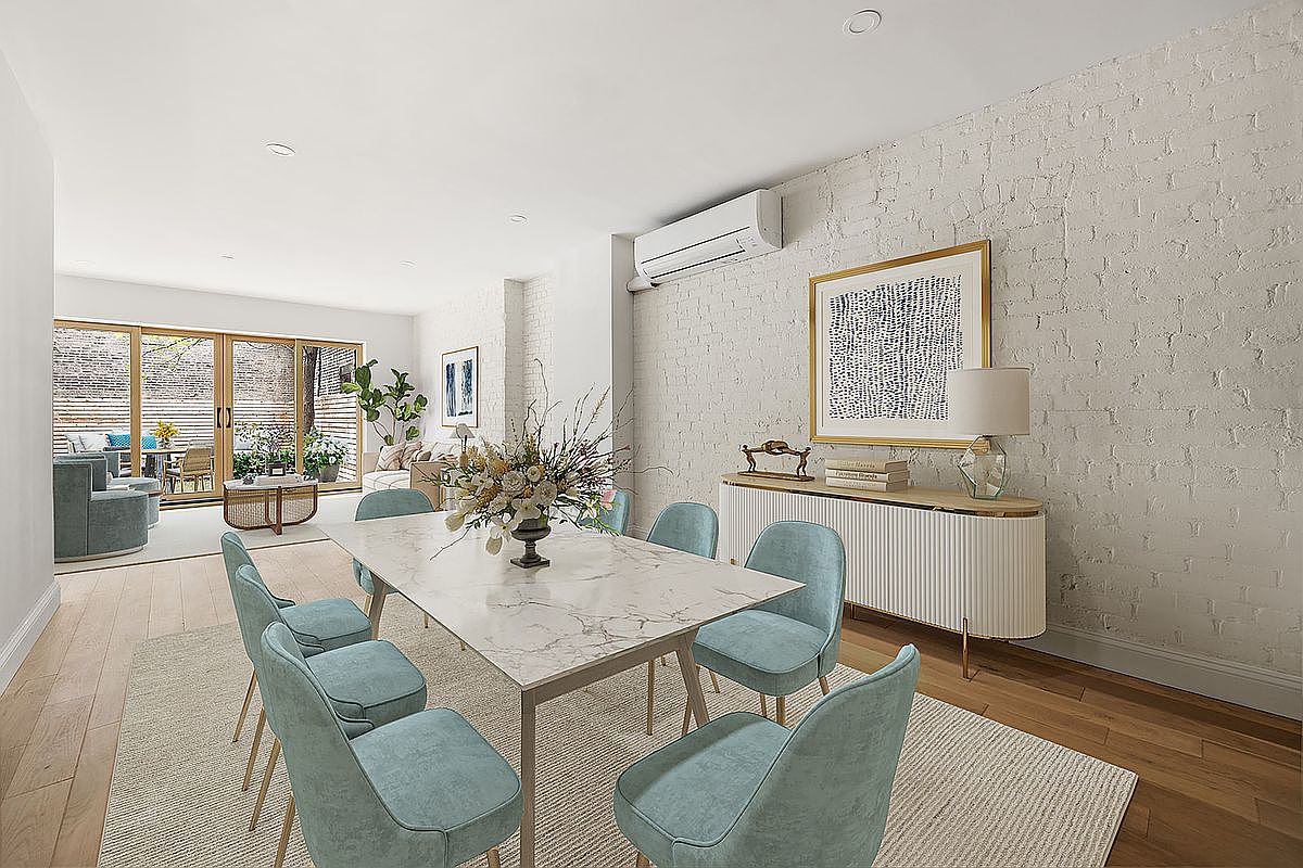 This is an interior shot of a dining room featuring a marble-top dining table surrounded by six light blue upholstered chairs. A modern white sideboard with a lamp and framed artwork sits against a white brick wall, and a large sliding glass door leads to an outdoor patio area. The room is well-lit and has a contemporary, inviting feel.