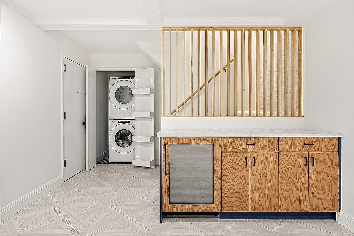 This interior shot showcases a well-organized laundry area featuring a stacked washer and dryer unit within a closet. Adjacent to the laundry closet is a stylish countertop with wooden cabinets and a built-in beverage cooler, all set against a backdrop of white walls and patterned flooring. The space is functional and aesthetically pleasing, blending utility with modern design.