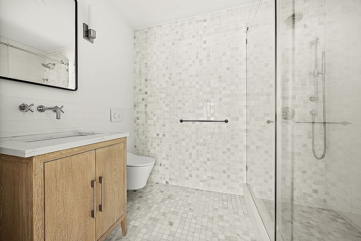 This is a well-lit bathroom featuring a modern vanity with a light wood finish and a white countertop. The walls and floor are tiled with small, light-colored mosaic tiles, creating a clean and bright aesthetic. A glass-enclosed shower is visible on the right, and a toilet is positioned next to the vanity.