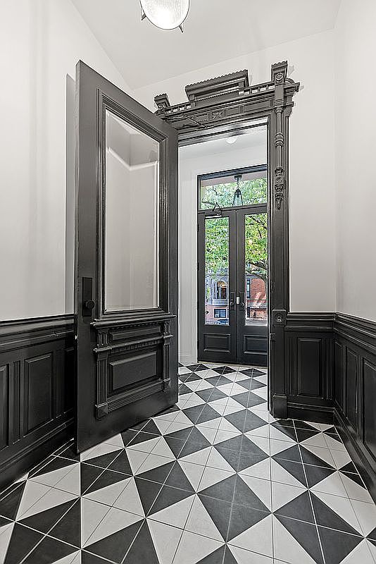 This interior shot showcases a grand entryway with striking black and white geometric tile flooring. An ornate, dark-painted door stands ajar, revealing a glimpse of the outdoors through a second set of doors. The walls feature wainscoting, adding to the classic and sophisticated aesthetic of the space.