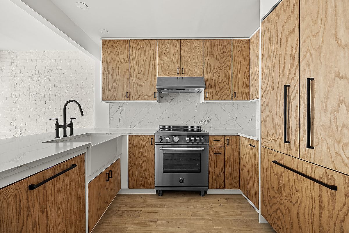 This is a well-lit kitchen featuring light wood cabinetry with black hardware, a white marble backsplash and countertops, and stainless steel appliances. The kitchen has a modern and minimalist design, with a focus on clean lines and natural materials. The flooring is light wood, complementing the cabinetry.