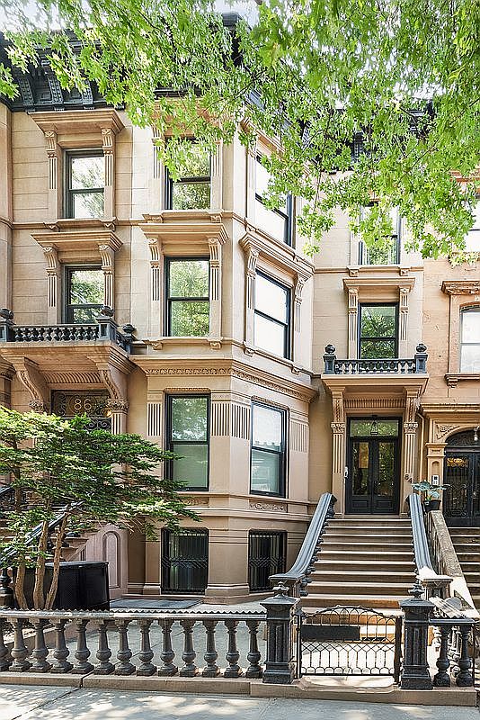 This is a front view of a classic brownstone building with ornate architectural details. The facade features multiple windows, decorative cornices, and a grand entrance with steps leading up to the front door. A wrought-iron fence and lush greenery add to the property's curb appeal, creating an inviting and sophisticated impression.