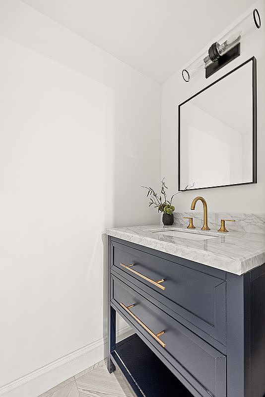 This is a well-lit bathroom featuring a navy blue vanity with gold hardware and a white marble countertop. A black-framed mirror hangs above the vanity, complemented by a modern light fixture. The overall design is stylish and clean, creating a sophisticated and inviting space.
