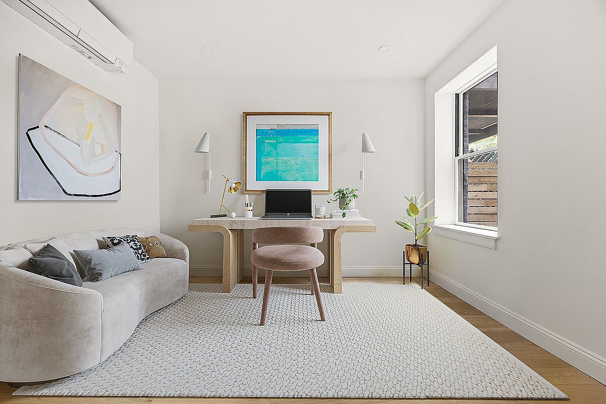 This is an interior shot of a bright and airy home office. The room features a modern desk with a laptop, a comfortable curved sofa with decorative pillows, and a large abstract painting on the wall. A window provides natural light, and a patterned rug covers the hardwood floor, creating a welcoming and productive workspace.