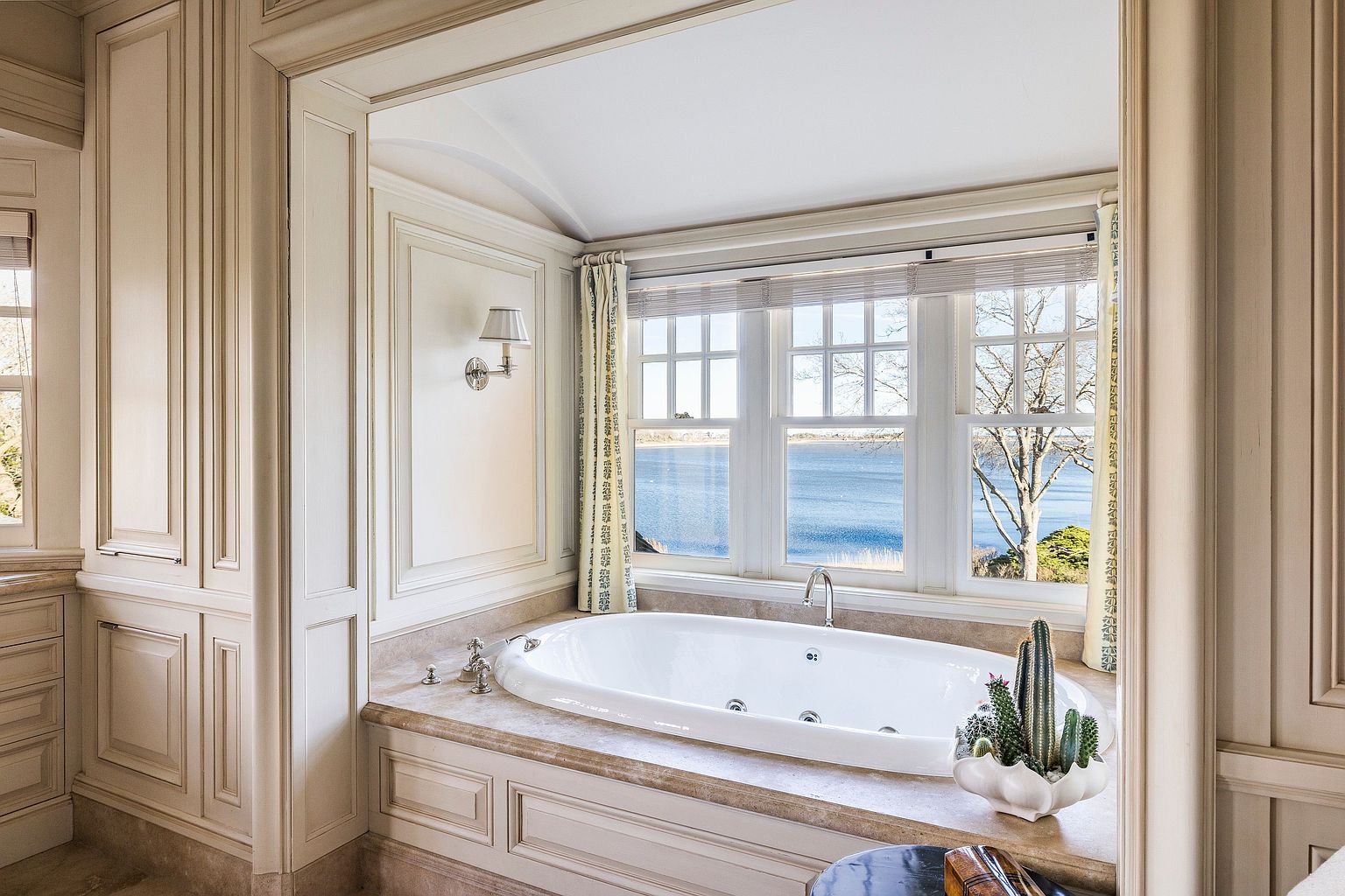 This is a luxurious primary bathroom featuring a large, oval-shaped soaking tub positioned in front of a window with a view of the water. The bathroom is decorated with neutral tones and detailed woodwork, creating a serene and elegant atmosphere. A potted cactus adds a touch of nature to the space.