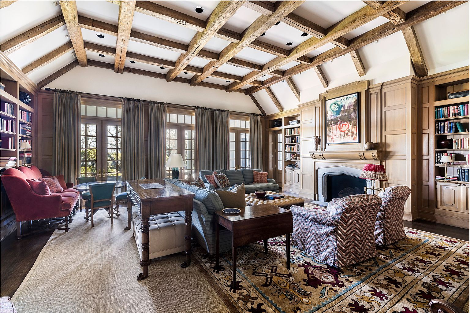 This is a spacious living room featuring a high, beamed ceiling and custom wood paneling. The room is furnished with comfortable seating arrangements, including sofas and armchairs, and is decorated with a mix of textures and patterns. The focal point is a fireplace with a decorative mantel, flanked by built-in bookshelves, creating a warm and inviting atmosphere.