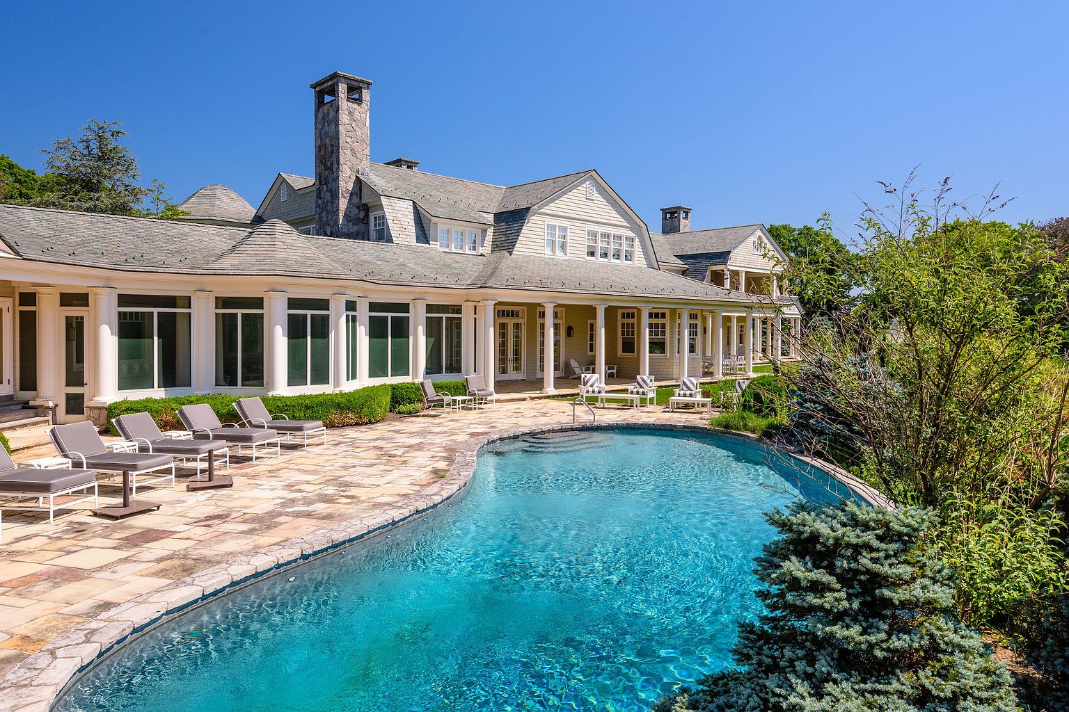 This image showcases a luxurious backyard featuring a large, freeform swimming pool with crystal clear blue water. The pool is surrounded by a stone patio with lounge chairs, perfect for relaxation. The house, with its elegant architecture, is visible in the background, complemented by lush landscaping that enhances the property's appeal.