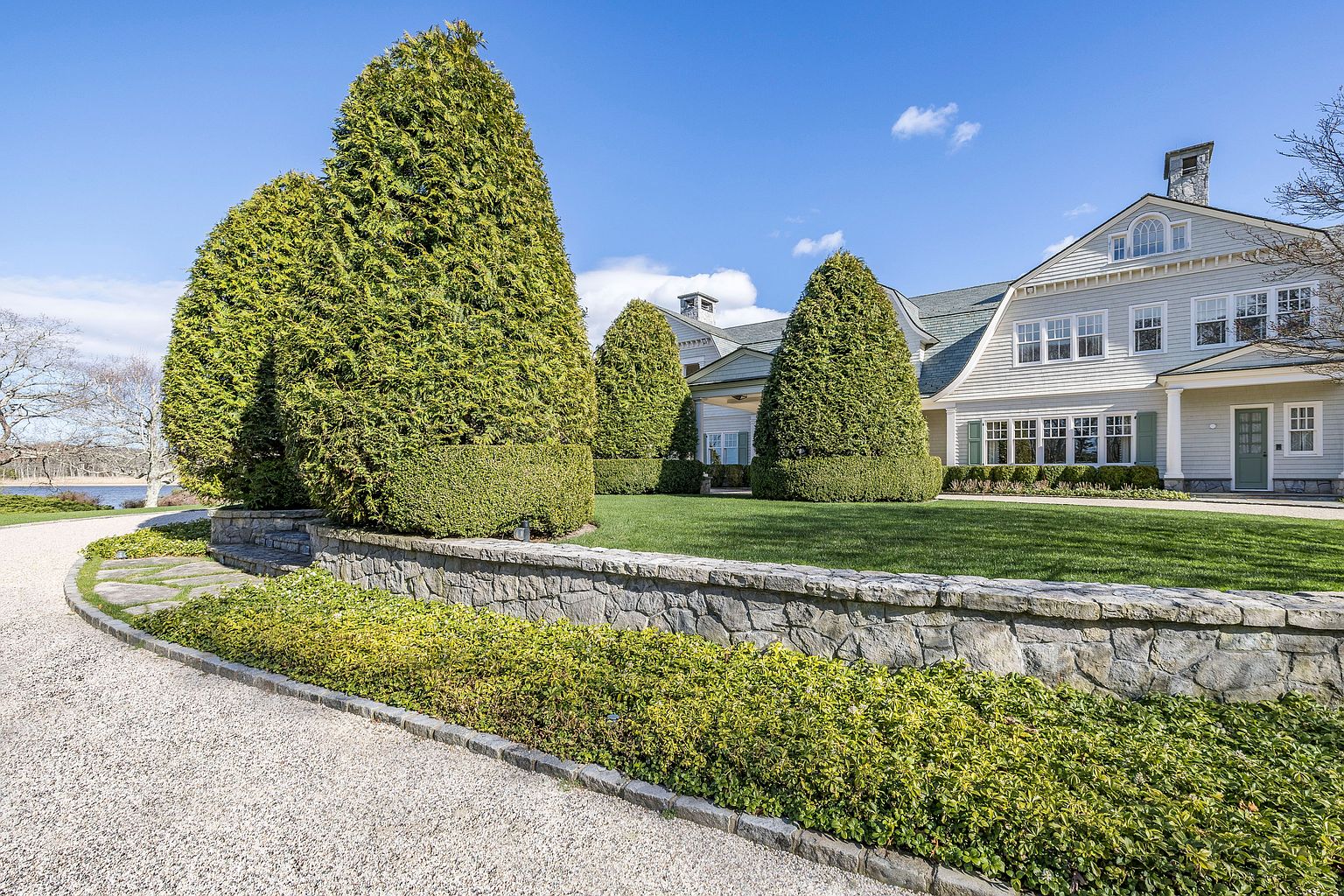 This image showcases the front exterior of a stately home with meticulously manicured landscaping. A stone wall lined with greenery adds to the property's curb appeal, while tall, sculpted trees frame the house. The residence features a classic architectural style with a light-colored facade and a well-maintained lawn, creating an inviting and luxurious impression.