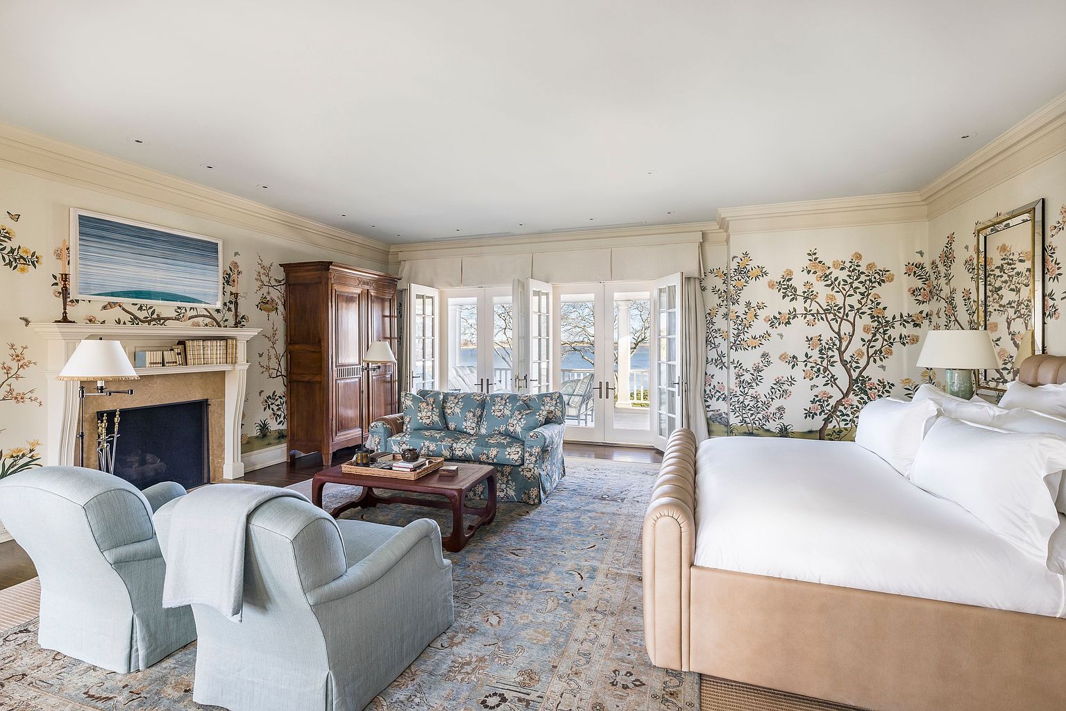This primary bedroom features a luxurious and serene atmosphere with floral wallpaper and a neutral color palette. The room includes a fireplace, a seating area with a floral sofa and light blue armchairs, and French doors leading to a balcony. A large bed with white linens and a tan upholstered frame is prominently displayed, complemented by an area rug that ties the room together.