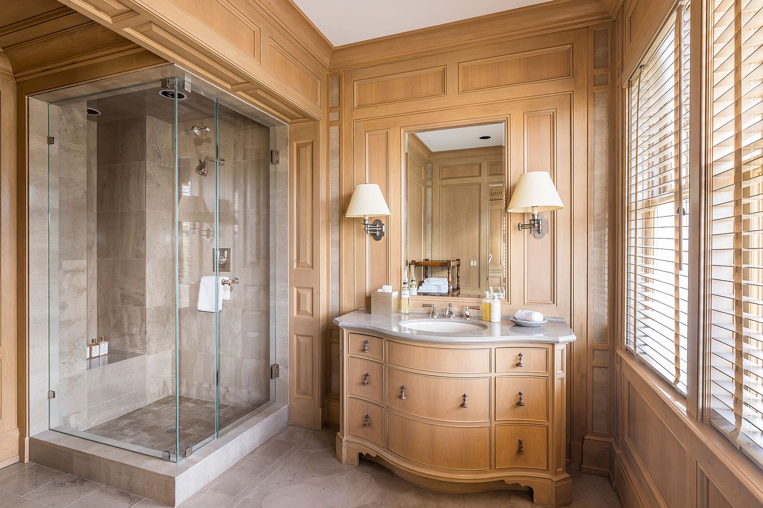 This is a well-appointed primary bathroom featuring wood paneling, a glass-enclosed shower, and a custom vanity with a marble countertop. The room is illuminated by sconces flanking a mirror above the vanity and natural light filtering through window blinds. The overall impression is one of classic elegance and luxury.